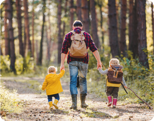 En la imagen se muestra un padre agarrandole la mano a sus dos hijos caminando por un bosque.